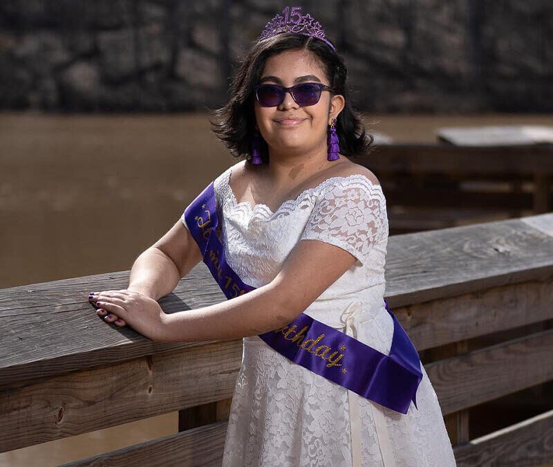 A girl in a white dress and purple sash is posing on a wooden bridge.