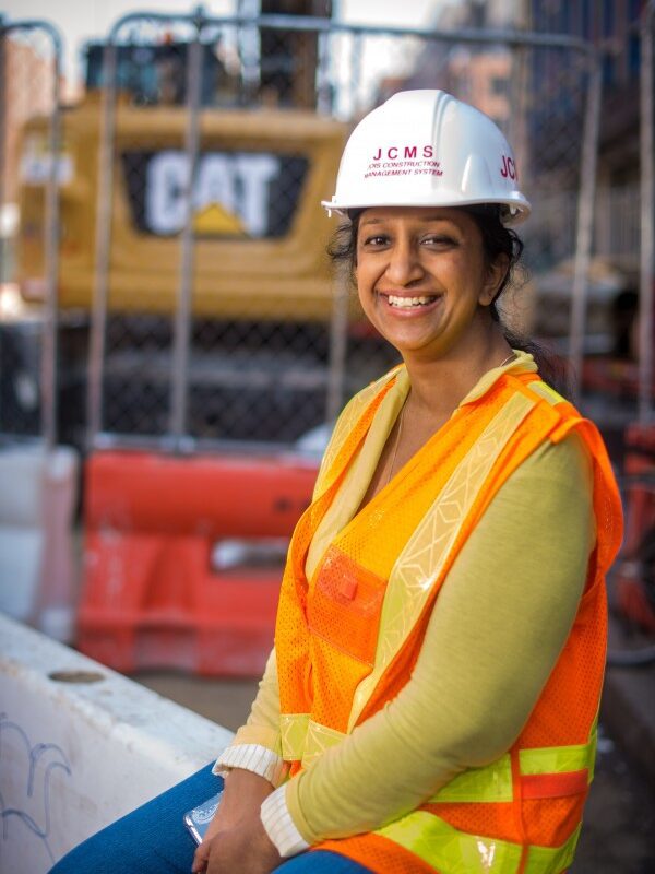 A woman wearing an orange vest and hard hat sits on a construction site.