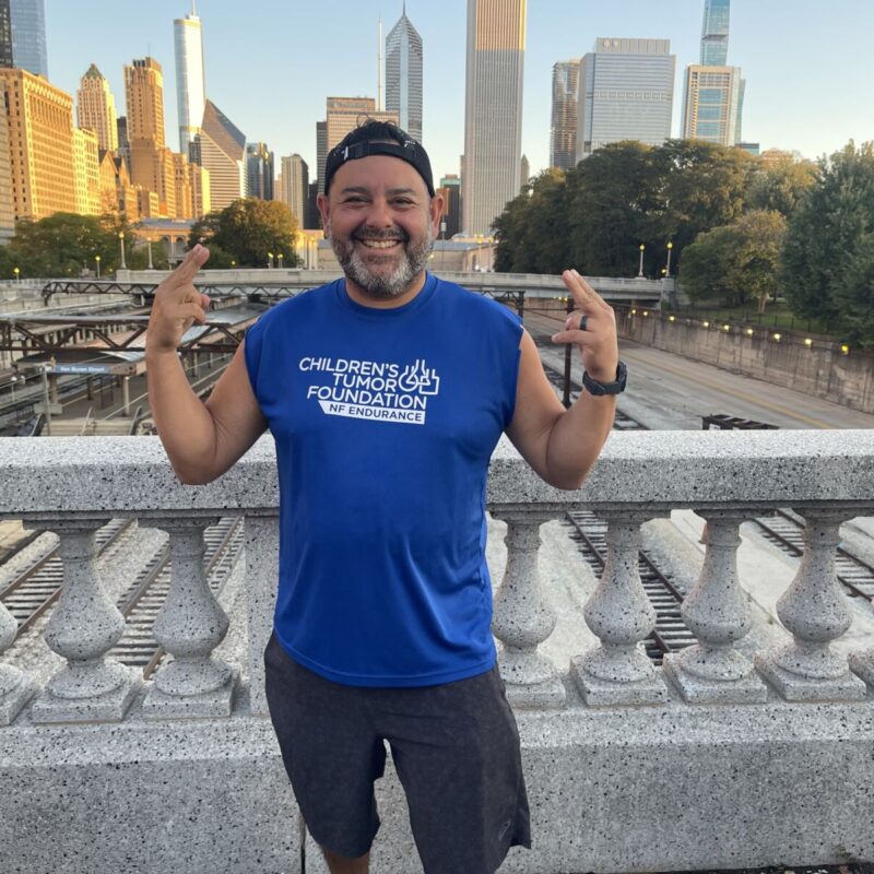 A man in a blue t-shirt smiles and makes a "rock on" gesture, standing on a bridge with a city skyline in the background during sunset.