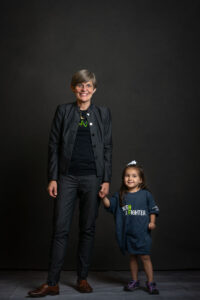 A woman in a suit stands holding hands with a small child wearing a "sunburn fighter" t-shirt, both smiling against a dark backdrop.