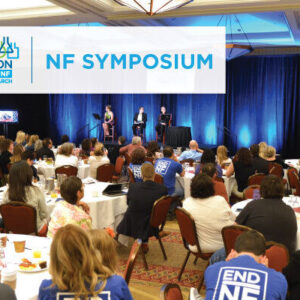 Participants seated at tables attending the Children's Tumor Foundation NF Symposium, with speakers on stage and a screen displaying a presentation in the background.