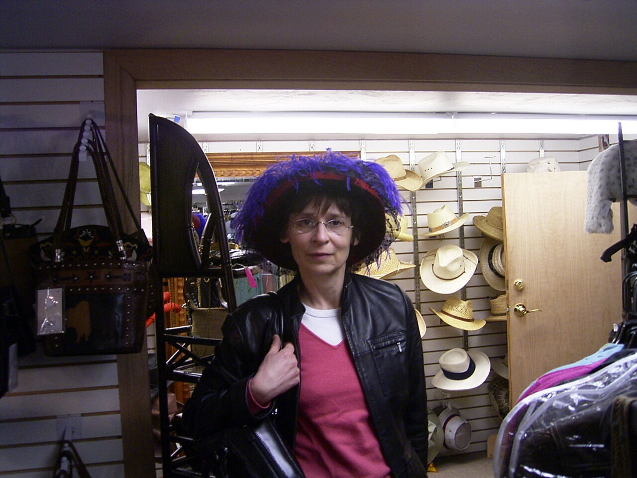A person in a black leather jacket and pink shirt stands indoors wearing a large purple and blue decorative hat, with various hats displayed on the shelves behind them.