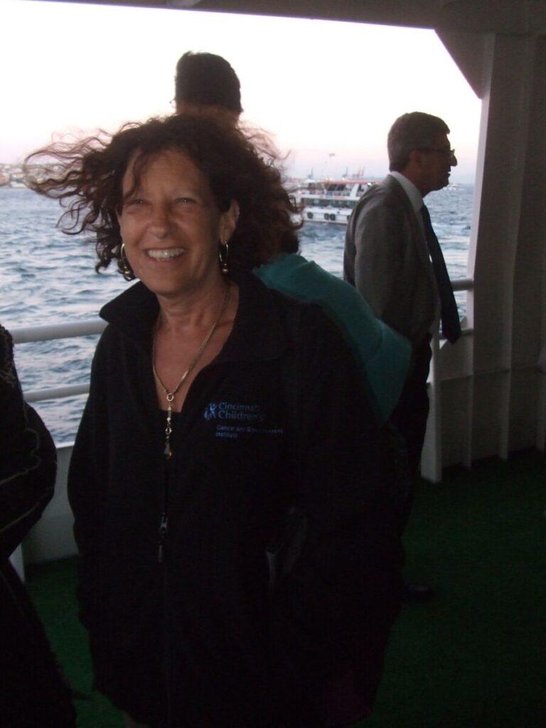 A woman with curly hair wearing a black jacket smiles while standing on a boat. Another man in a suit stands in the background by the railing, with water and another boat visible behind them.