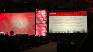 A conference stage with large screens displaying the event schedule, surrounded by attendees in a dimly lit hall.