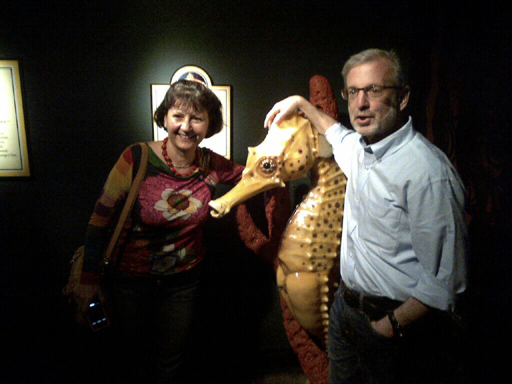 A woman and a man stand on either side of a large seahorse sculpture. The woman is smiling, and the man has his hand on the seahorse's head. They are in a dimly lit room.