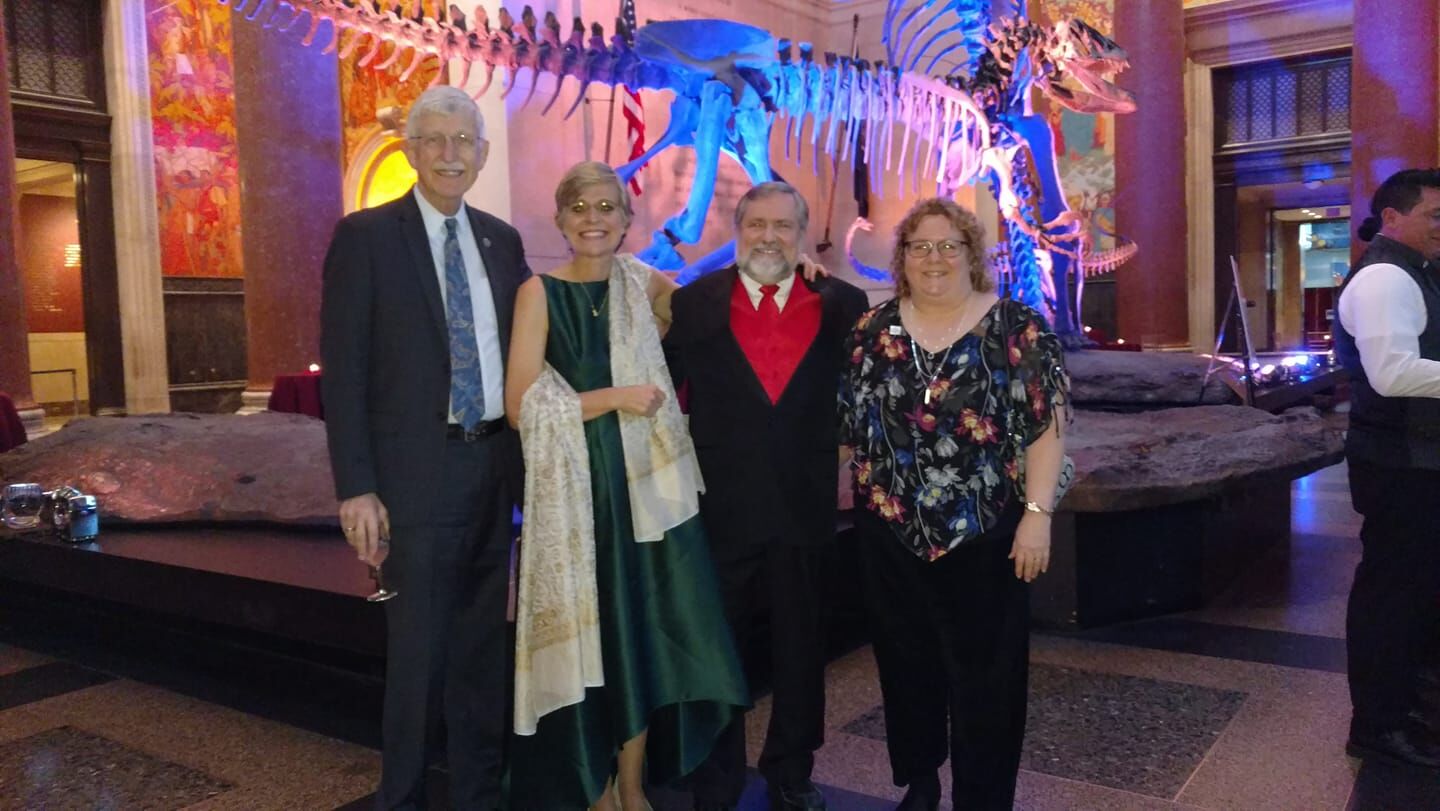 Four people pose in front of a dinosaur skeleton display in a museum. Two of them wear formal attire, and the other two are dressed casually. The background features museum decor and lighting.