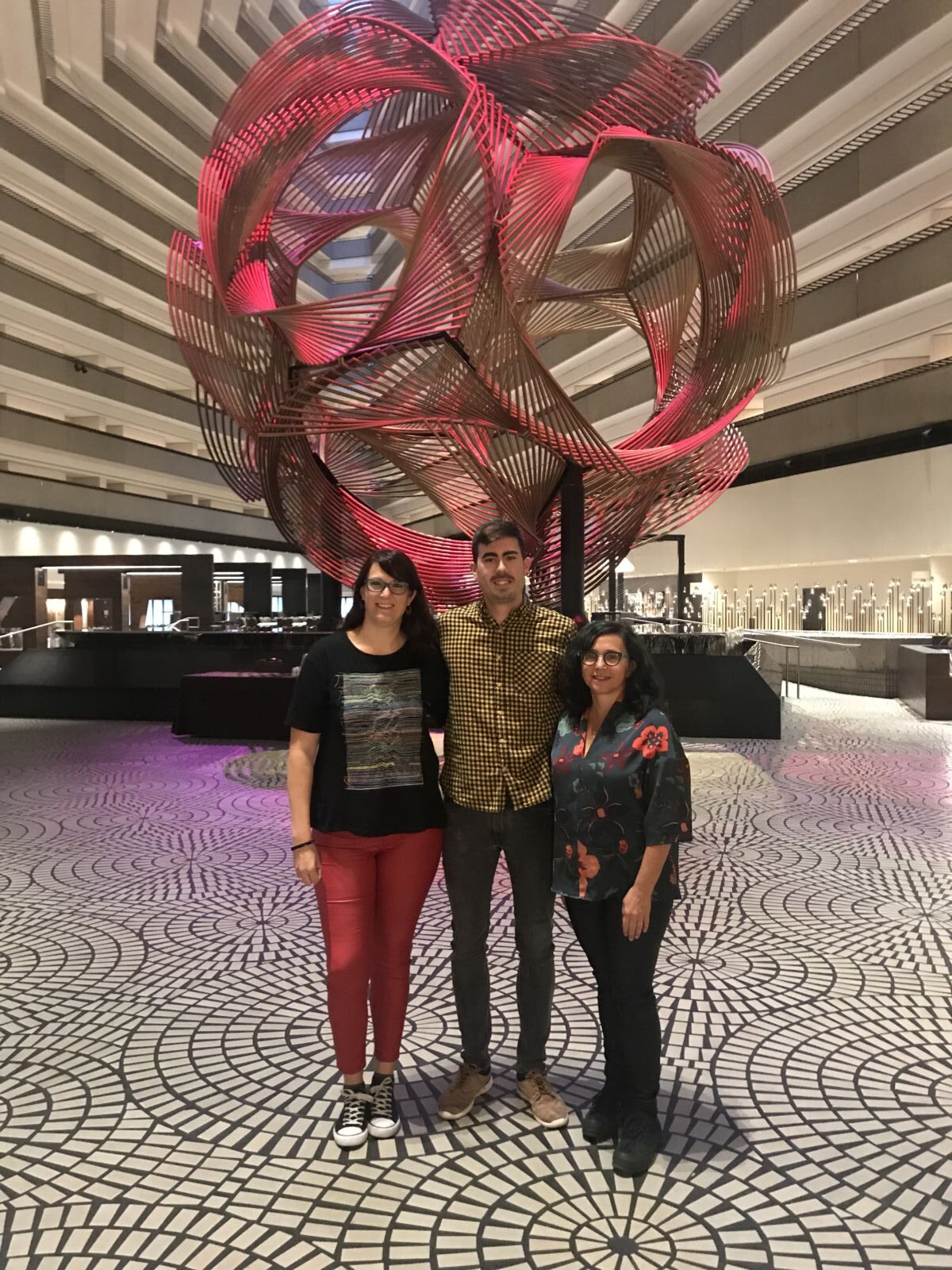 Three people stand together in a patterned lobby with a large, abstract red sculpture in the background.