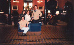 Four people are posing inside a grand room with red walls, chandeliers, and ornate furnishings. Two are sitting, and two are standing behind a blue velvet couch.
