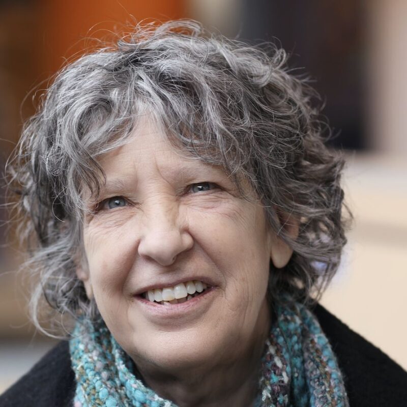 A close-up of an older woman with curly gray hair and a scarf, smiling while looking slightly to the side. The background is softly blurred.