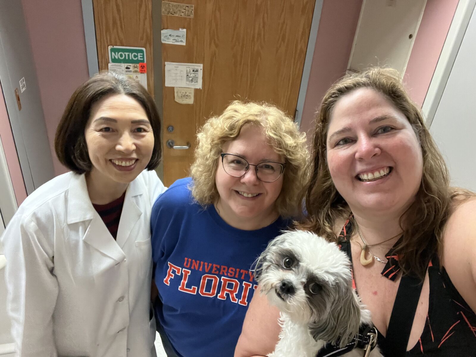 Three women and a small dog pose for a photo inside a room with a wooden door in the background. One woman wears a white lab coat, another has a University of Florida shirt, and the third holds the dog.