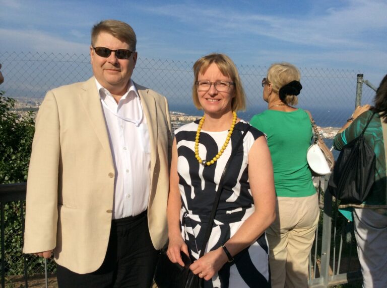 A man and woman stand outdoors in front of a chain-link fence with a coastal city in the background. The man wears sunglasses and a beige jacket, the woman wears glasses and a black-and-white dress.