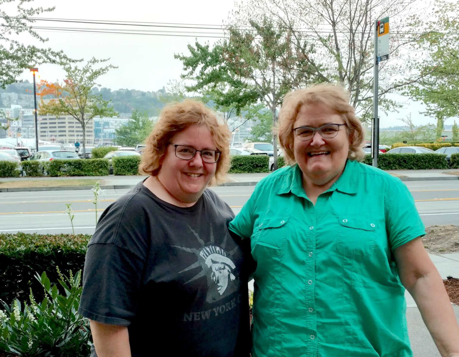 Two smiling women stand outdoors by a tree-lined street and parking lot, with one wearing a gray Statue of Liberty T-shirt and the other a green button-up shirt.