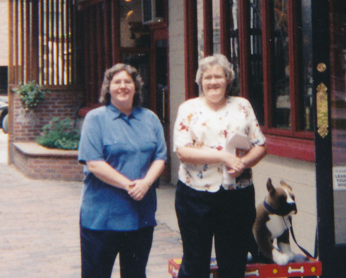 Two women standing side by side on a brick walkway in front of a building with glass windows and a red door; an artificial dog figure is placed on the ground beside them.