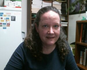 A woman with curly hair is seated, looking at the camera. Behind her are shelves with books and a white refrigerator with colorful magnets.