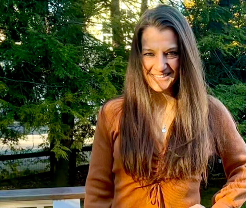 A person with long brown hair, wearing an orange long-sleeve top, stands outdoors in front of a forest-like background, smiling at the camera.
