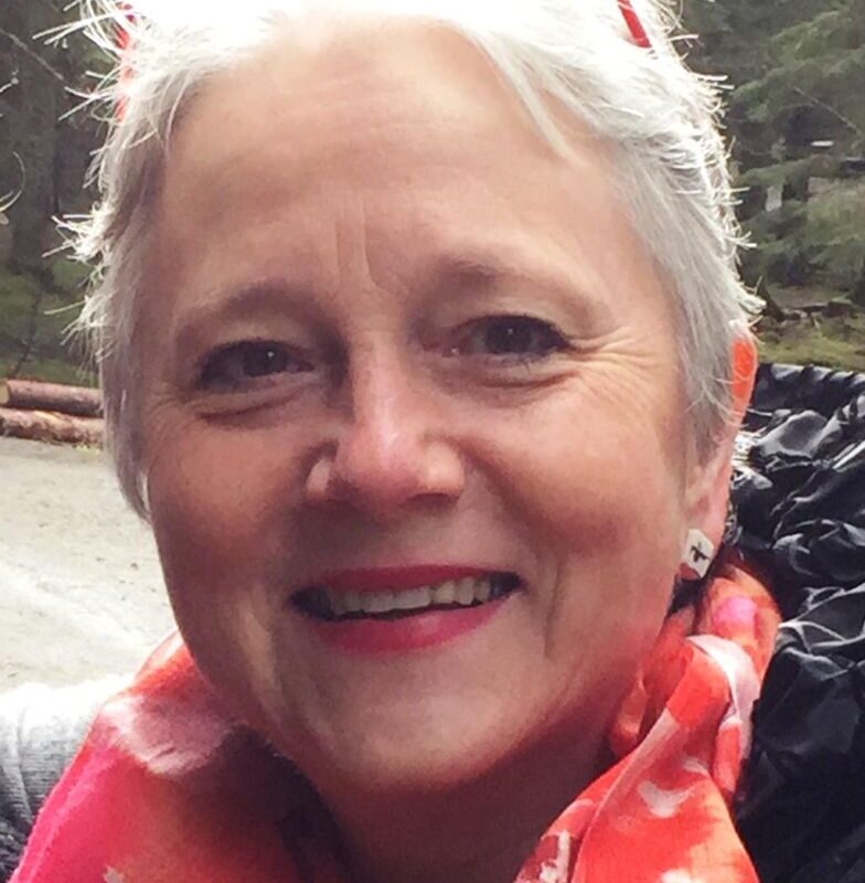 A smiling woman with short gray hair and orange-framed glasses on her head, wearing a pink patterned scarf and a black jacket. The background shows an outdoor, wooded area.