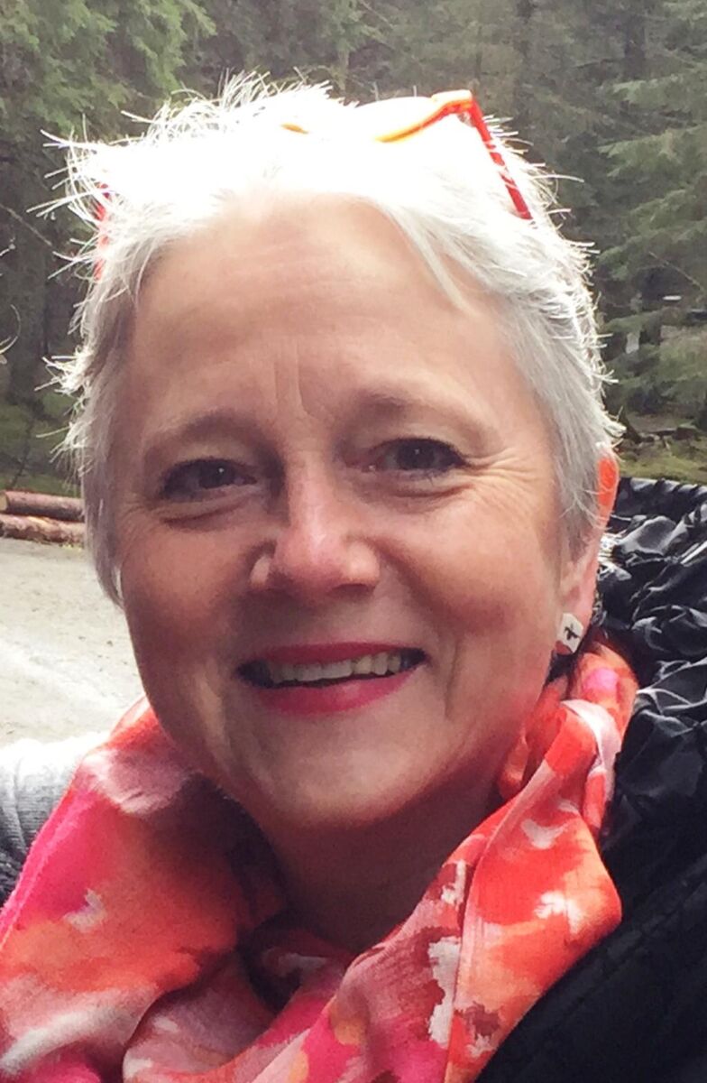 A smiling woman with short gray hair and orange-framed glasses on her head, wearing a pink patterned scarf and a black jacket. The background shows an outdoor, wooded area.