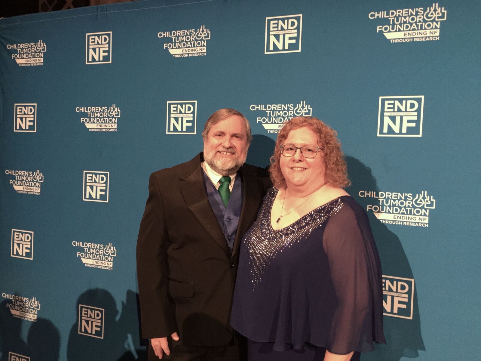 A man in a dark suit and a woman in a blue dress pose together in front of a blue backdrop with "END NF" and "Children's Tumor Foundation" logos.