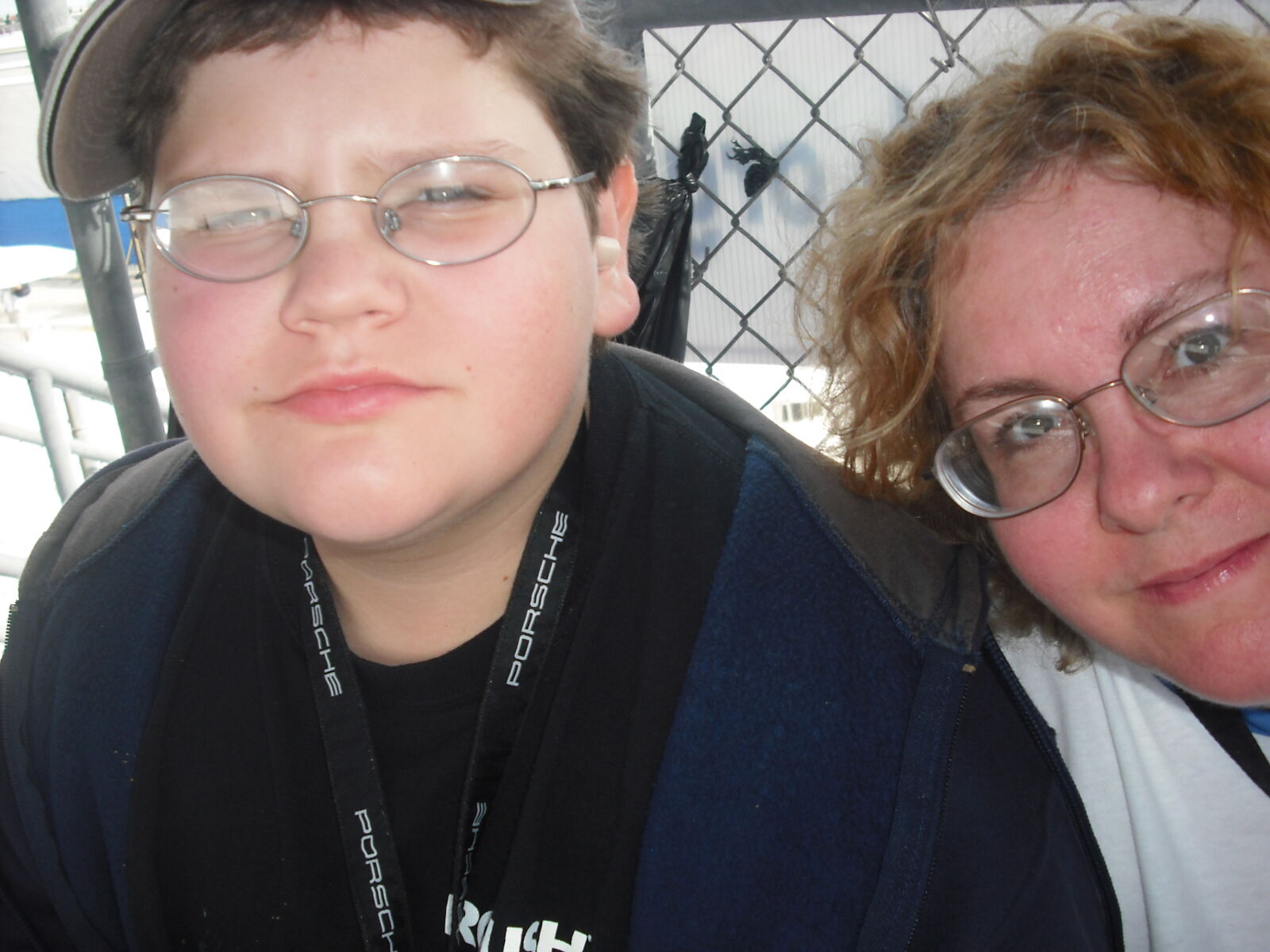 A young boy and a woman, both wearing glasses, smile at the camera while standing near a chain-link fence.