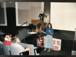 A woman is giving a presentation in an auditorium. She stands next to a podium, and a banner in front reads "IN" followed by "RACE TO THE FUTURE." An audience is seated watching her.