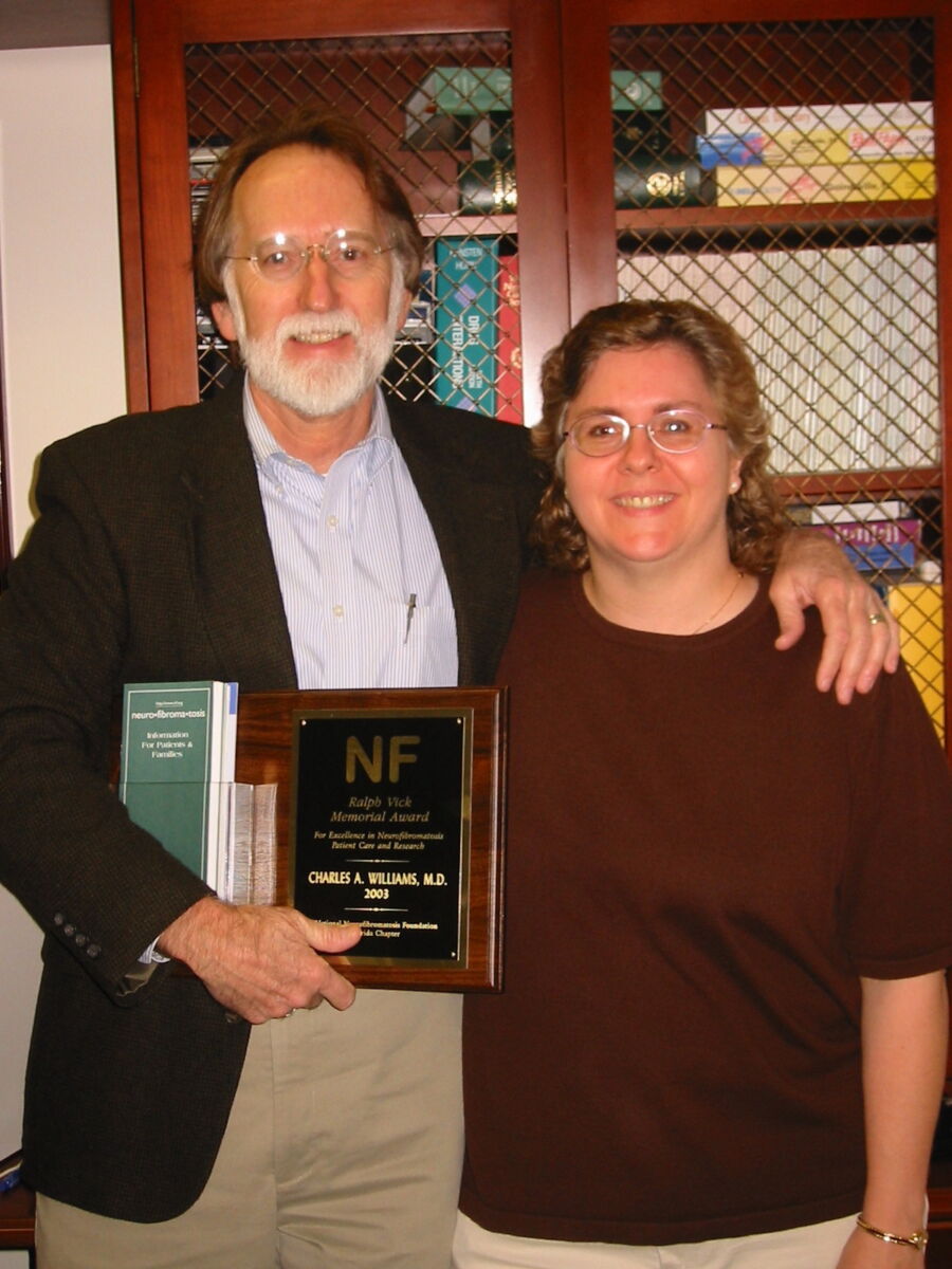Two people stand together smiling. The person on the left holds an award plaque and a book. They are in front of a wooden bookshelf.