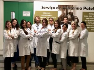 A group of eleven people, wearing white lab coats, stand together in front of a sign that reads "Serviço de Anatomia Patológica." They pose with arms crossed, smiling at the camera.