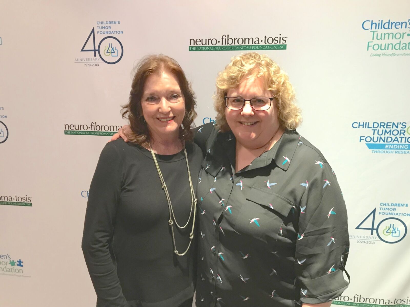 Two people standing in front of a backdrop with logos of the Children's Tumor Foundation and neurofibromatosis organizations. Both are smiling at the camera.