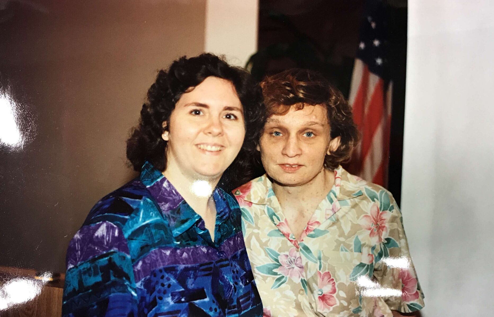 Two people are standing close together and smiling. They are both wearing patterned shirts. An American flag is partially visible in the background.