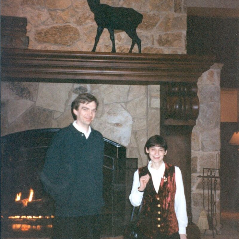 Two people pose in front of a stone fireplace with a lit fire, beneath a mantle displaying a decorative deer silhouette.