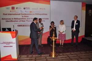 A man lights a ceremonial lamp while four individuals stand by at the First International Conference on RASopathies in Asia. A banner with conference details is displayed in the background.