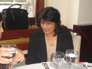 A woman in a beige top and black blazer is sitting at a table with a white tablecloth, engaged in conversation. There are glasses of water and a black handbag on the table. Photo date is November 1, 2009.