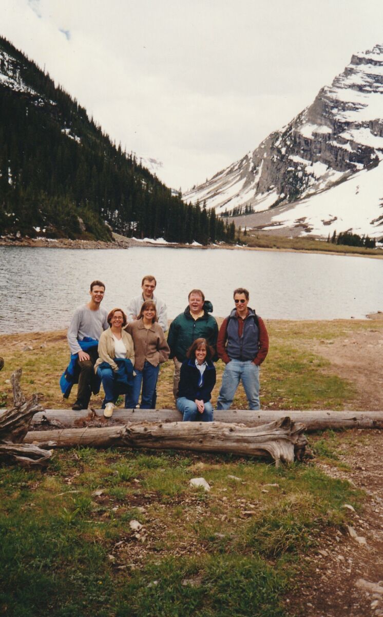 A group of seven people posing on a grassy area by a log near a mountain lake with snow-covered mountains and pine trees in the background.