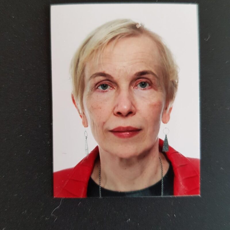 A portrait photograph of an older woman with short blonde hair, wearing a red jacket, black top, and dangling earrings, against a plain background.