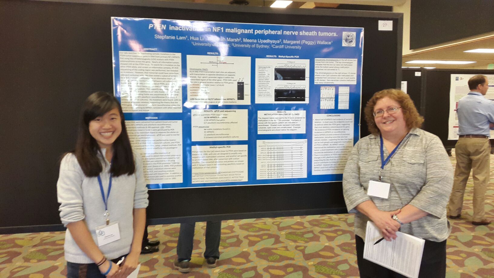 Two individuals stand in front of a scientific poster presentation titled "PTEN Inactivation in NF1 Malignant Peripheral Nerve Sheath Tumors." The poster includes text, charts, and images.