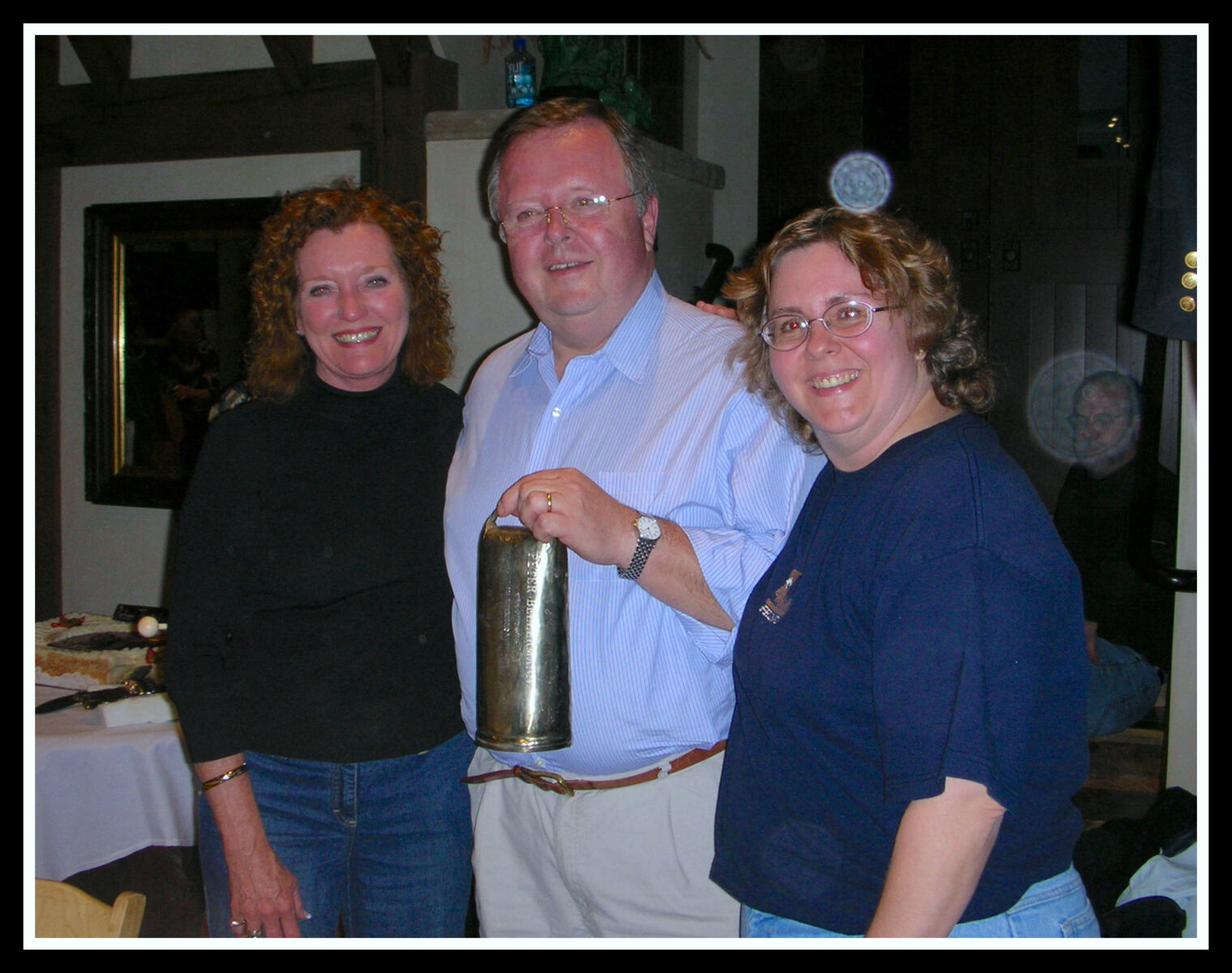 Three people stand indoors, smiling at the camera. The person in the middle holds a shiny metallic object. A table is visible in the background.