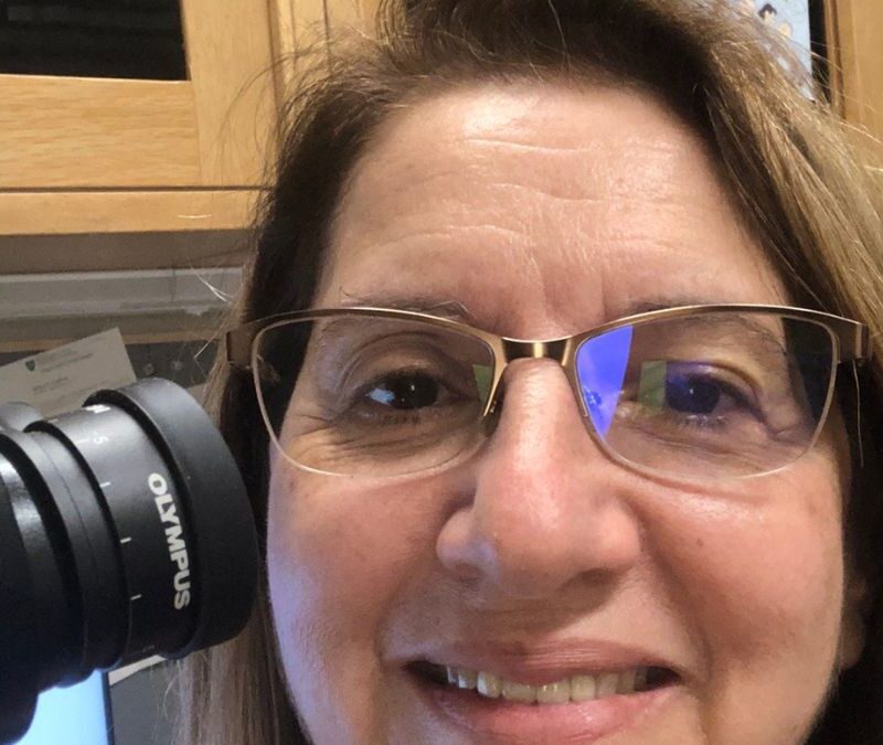 A person with glasses smiles at the camera while looking through an Olympus microscope.