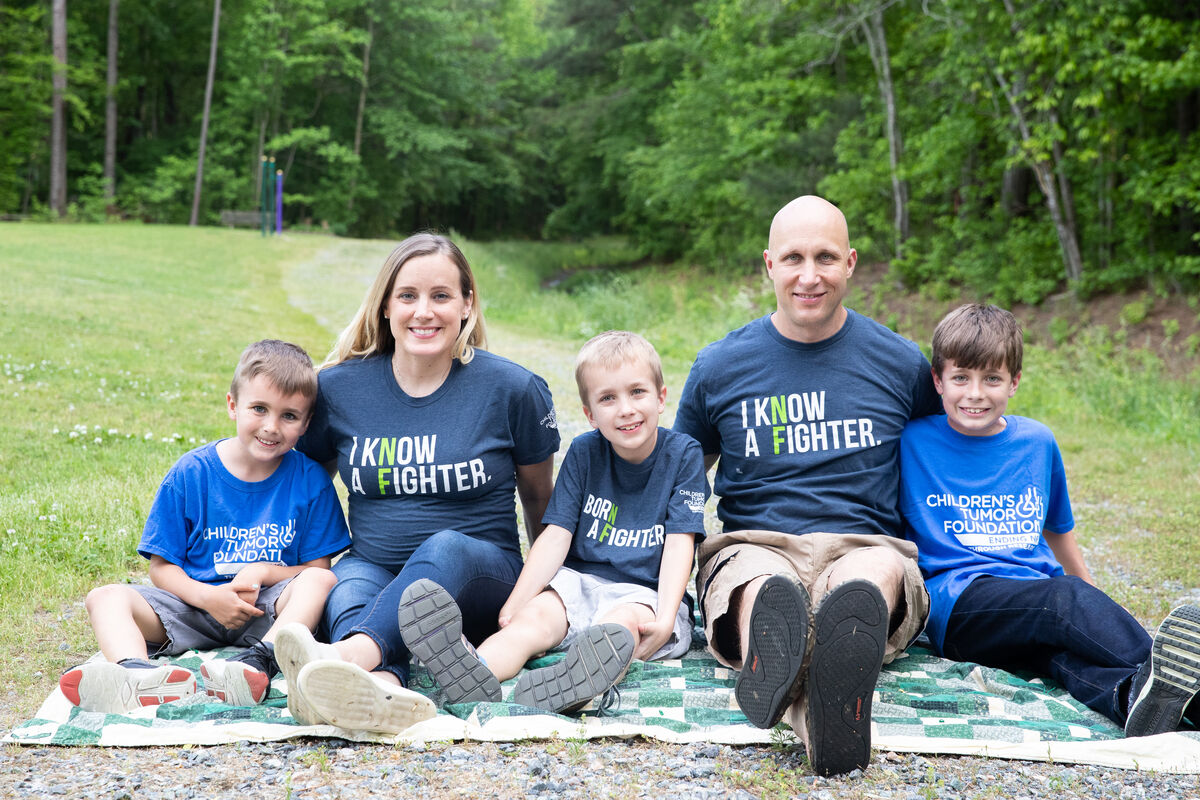 A family of five sits on a picnic blanket outdoors. The parents and one child wear shirts that say "I Know A Fighter;" the other two children wear shirts that say "Children's Tumor Foundation.