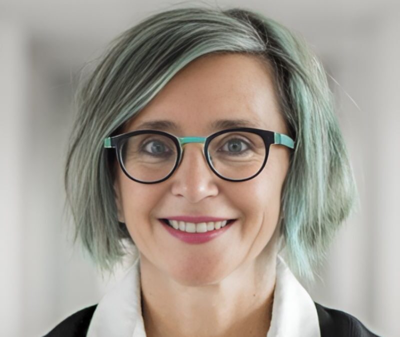 A person with short, gray and green hair, wearing glasses and a white-collared shirt, smiles at the camera.