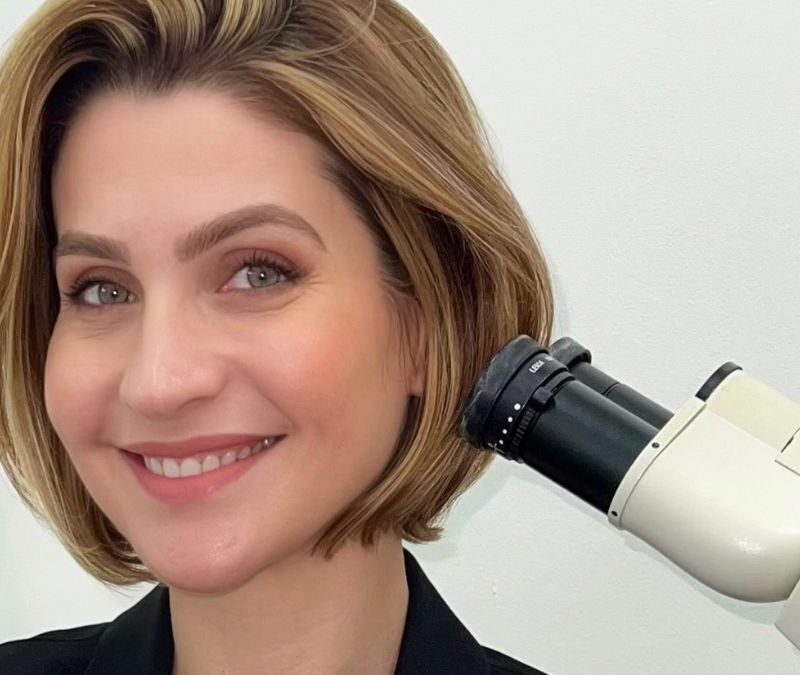 A person with short hair smiles while sitting next to a microscope.