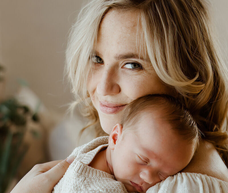 A woman with blonde hair is holding a sleeping baby against her chest. She is smiling softly at the camera, and there is a plant in the blurred background.