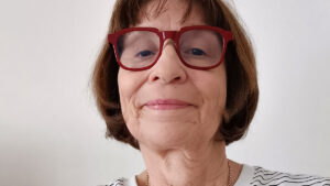 A woman with short brown hair and red glasses, wearing a white shirt with black stripes, looks at the camera against a plain background.
