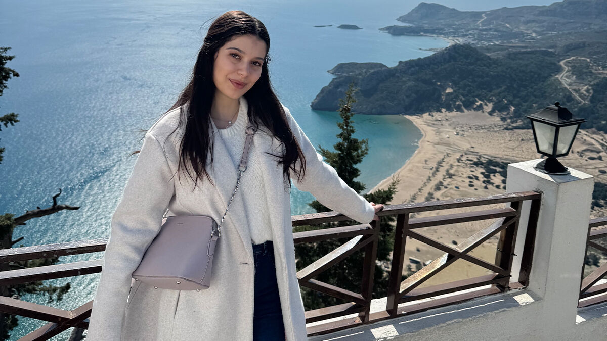 A woman stands by a wooden railing overlooking a coastal landscape with mountains and a sandy beach in the background. She is wearing a light coat and carrying a small bag.