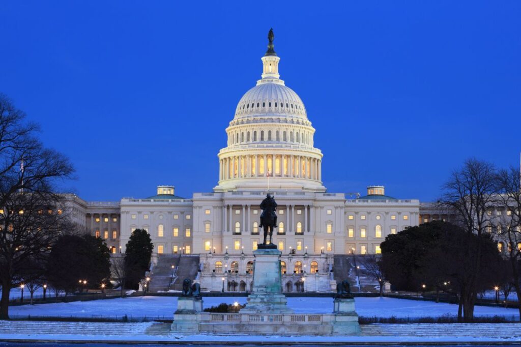 The U.S. Capitol building at dusk, illuminated against a deep blue sky, with the Ulysses S. Grant Memorial statue in the foreground.