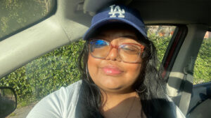 A person wearing large glasses and a blue baseball cap with an "LA" logo is sitting in a car, smiling slightly. Sunlight illuminates their face.