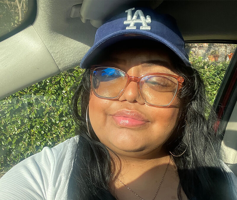 A person wearing large glasses and a blue baseball cap with an "LA" logo is sitting in a car, smiling slightly. Sunlight illuminates their face.
