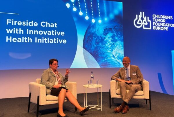 Two people seated on stage in discussion during the "Fireside Chat with Innovative Health Initiative" at a Children's Tumor Foundation Europe event. A screen behind them displays the event title.