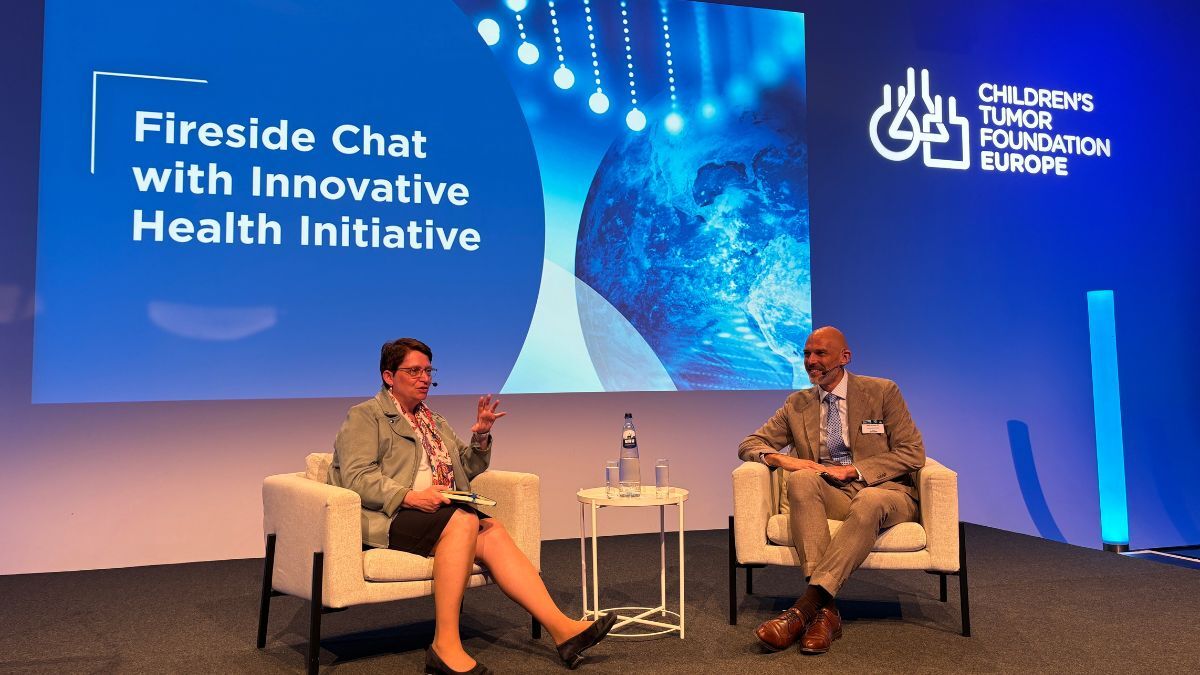 Two people seated on stage in discussion during the "Fireside Chat with Innovative Health Initiative" at a Children's Tumor Foundation Europe event. A screen behind them displays the event title.