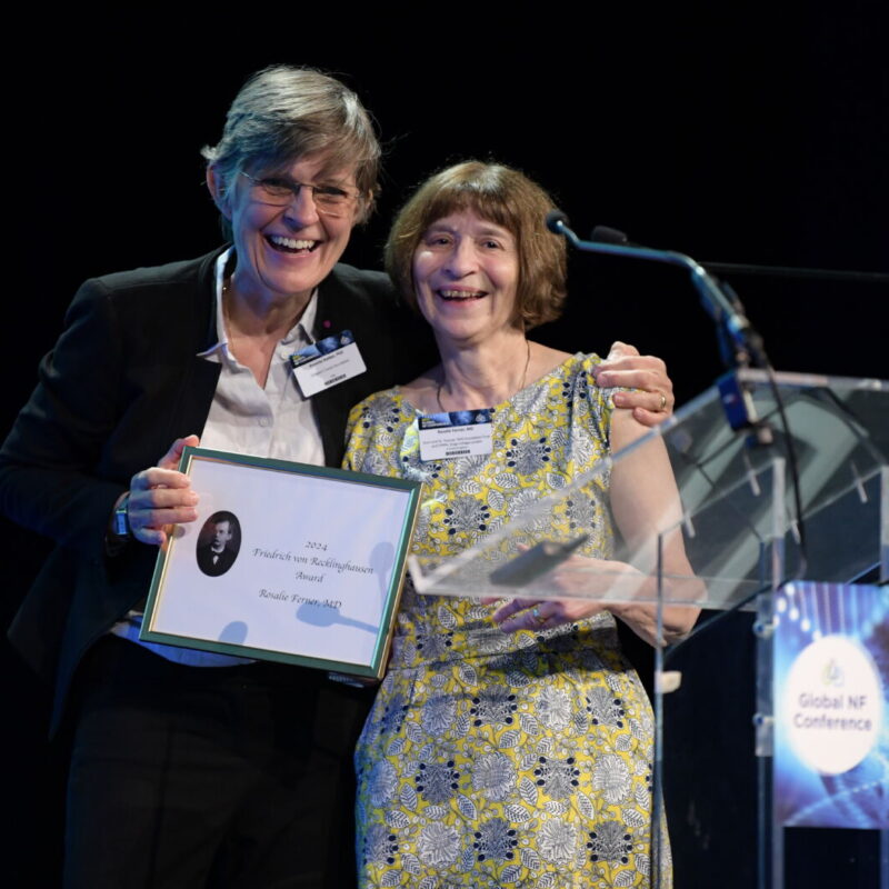 Two individuals on stage smile while holding an award. They stand beside a podium with the text "Global NF Conference." One person holds a plaque with the words "2014 Friedrich von Recklinghausen Award.