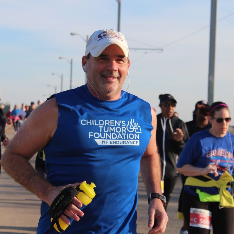 A man in a blue "Children's Tumor Foundation NF Endurance" shirt runs in a race, holding a yellow water bottle. Other runners are visible in the background.