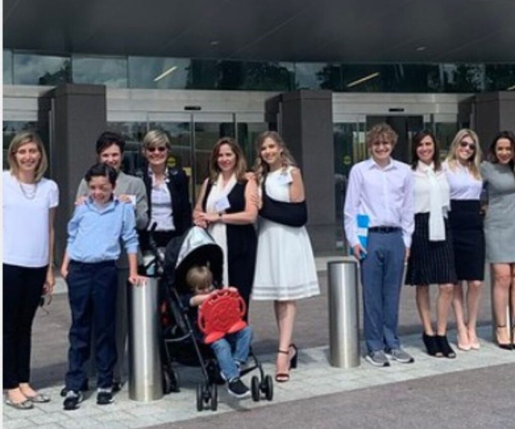 A group of people, including children and adults, are standing outdoors. One person is pushing a stroller with a young child, and another person has an arm in a sling. They are posing in front of a building.